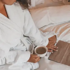 אישה מצולמת מלמעלה שותה קפה על מיטה של מלון